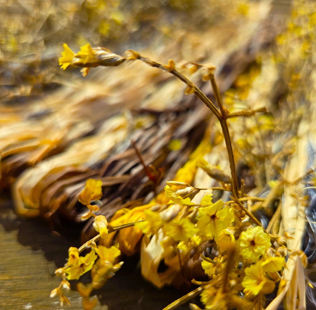 A photo of a detail of a woven artwork with twigs of small yellow flowers in the foreground, blurring to strips of brown, sandy beige, and speckled yellow in the middle- to background. There are some blue threads appearing on the lower right of the image. 